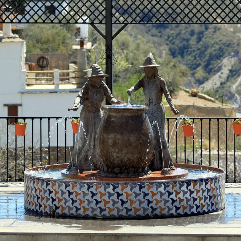 Fuente ornamental con dos figuras femeninas vertiendo agua desde un gran caldero sobre un pilón de azulejos, con un balcón de rejería, casas blancas y montañas al fondo.