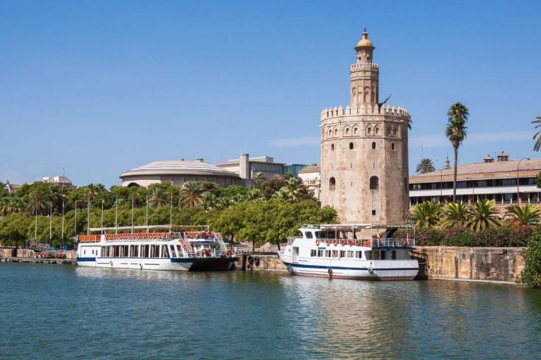vista de la torre del oro junto al río guadalquivir con barcos atracados en primer plano y cielo azul despejado.