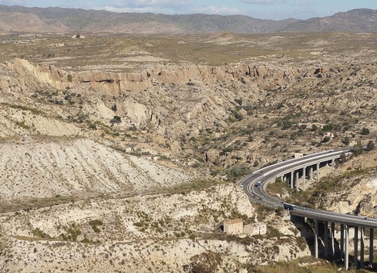 paisaje árido con estratos yesíferos erosionados y un viaducto de carretera que serpentea sobre un cañón rocoso