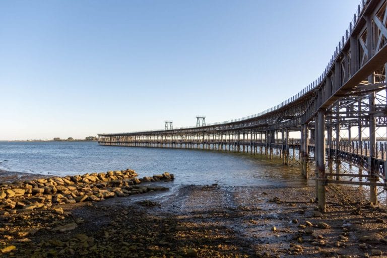 Panorámica del río tinto en Huelva con un muelle de hierro apoyado sobre pilares y rocas en primer plano.