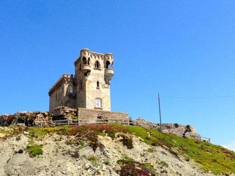 Fuerte blanco de forma cilíndrica y almenado, el Castillo de Santa Catalina, encajado en un promontorio rocoso junto al mar, con el horizonte de la costa y el cielo azul al fondo.