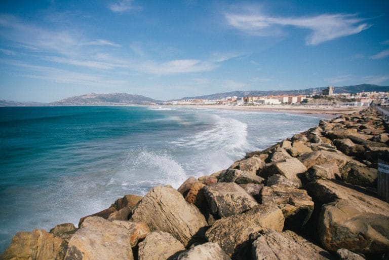 Playa de Los Lances en Tarifa: rompientes sobre la escollera de rocas, oleaje suave y la línea urbana del pueblo costero al fondo, bajo un cielo parcialmente nublado.