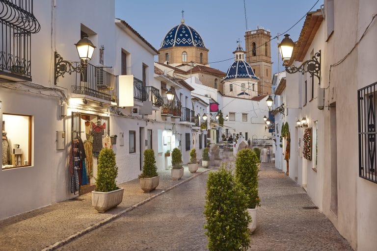 Calle empedrada y estrecha en el casco antiguo de Altea, con casas blancas, faroles y la cúpula azul de la iglesia de fondo