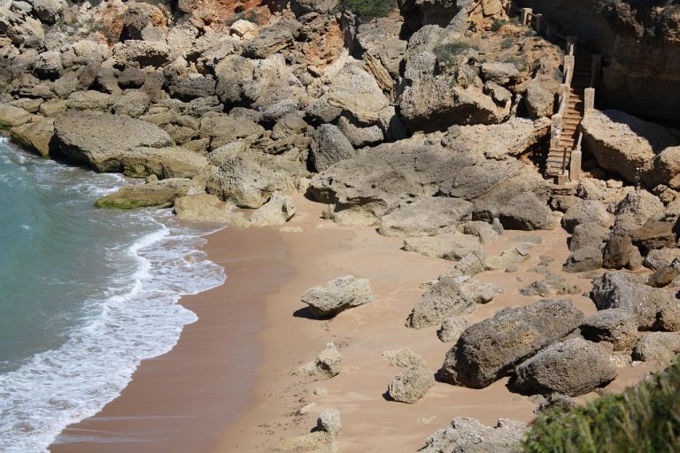 Escaleras de piedra que descienden a una pequeña cala rocosa en Conil de la Frontera, con rocas dispersas en la arena y suaves olas del Atlántico rompiendo en la orilla.