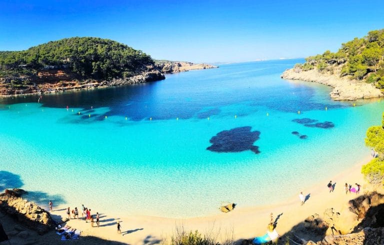 Vista panorámica de Cala Salada en Ibiza con aguas turquesas, playa de arena y rocas con gente disfrutando del día