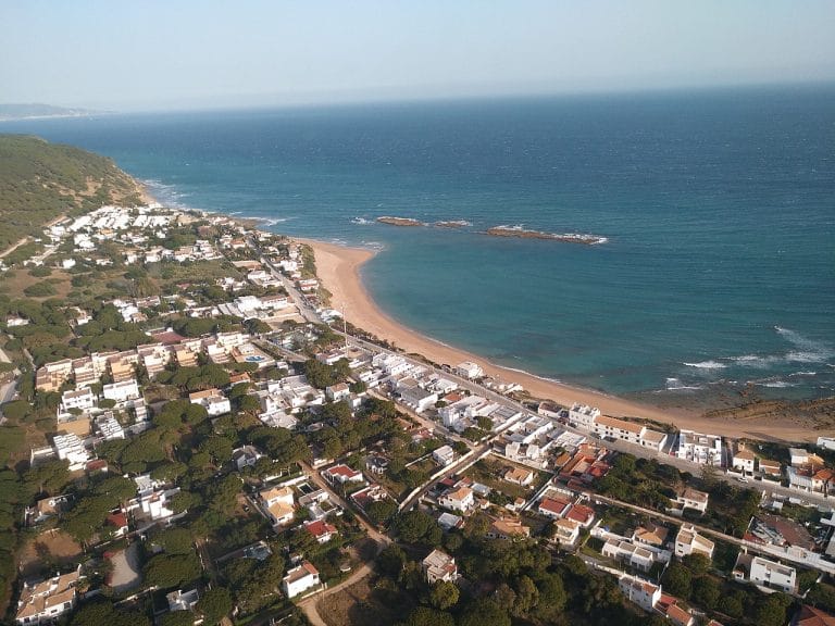 Vista aérea de Los Caños de Meca en Cádiz: playa dorada bordeada por casas blancas y marisma, con el océano Atlántico al fondo