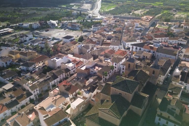 Fotografía aérea que muestra un entramado de calles estrechas y tejados de teja en una población de estilo tradicional, con casas agrupadas alrededor de la iglesia y rodeadas de campos.