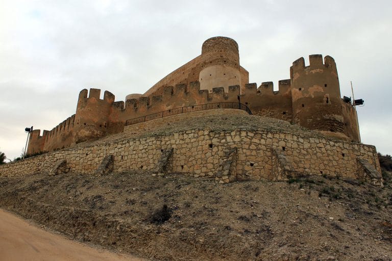 Vista frontal en contrapicado del castillo de Biar (Alicante), mostrando sus robustas murallas almenadas y torres cilíndricas sobre un basamento de mampostería, con un cielo nublado de fondo.