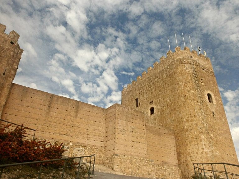 Vista del castillo de jumilla con sus murallas de piedra y torre cuadrada bajo un cielo parcialmente nublado.