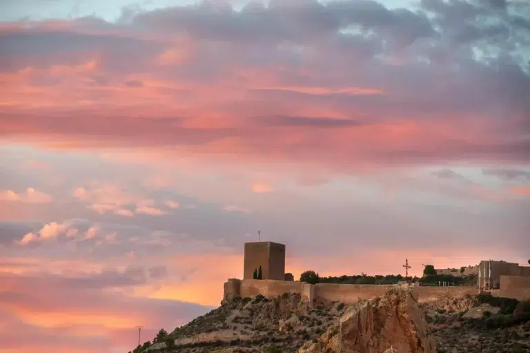 Atardecer sobre el castillo de Lorca con murallas de piedra y un cielo teñido de tonos rosados y naranjas.