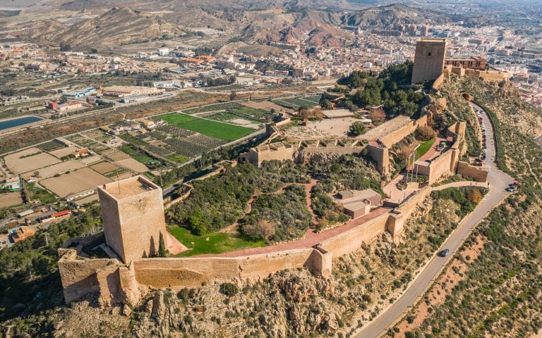 Vista aérea del castillo de Lorca sobre un cerro con murallas y torres y la ciudad y campos agrícolas al fondo.