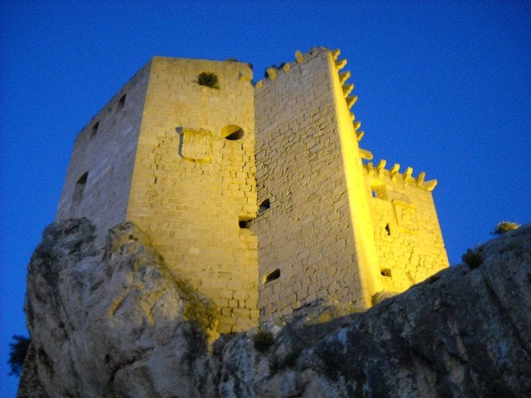 Fachada iluminada del castillo de Mula al anochecer, sobre un peñasco rocoso con luminosos tonos dorados contra un cielo azul profundo.