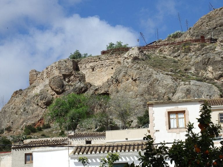 Ruinas del antiguo castillo de Purchena sobre un cerro rocoso, con una barandilla de madera en la parte superior; al pie del cerro se ven casas encaladas con tejados de teja y naranjos en primer plano, bajo un cielo azul con nubes dispersas.