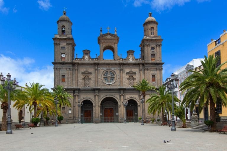 Fachada de la Catedral de Canarias en Las Palmas de Gran Canaria, con palmeras y cielo azul despejado.
