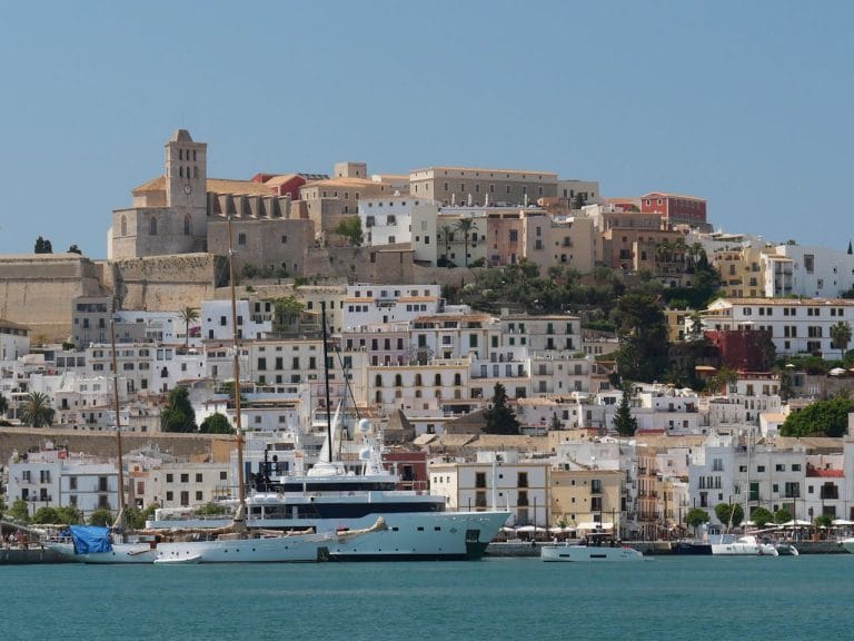 Vista del castillo de Ibiza con edificios históricos y modernos, puerto con barcos y yates bajo un cielo despejado