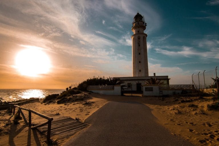 Faro de Trafalgar junto al mar al atardecer, con el sol poniéndose sobre el Atlántico y sus reflejos dorados sobre la arena