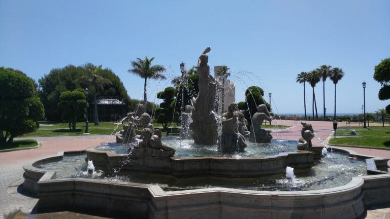 Fuente ornamental con estatuas en un parque costero de España, con palmeras y el mar de fondo bajo un cielo azul.