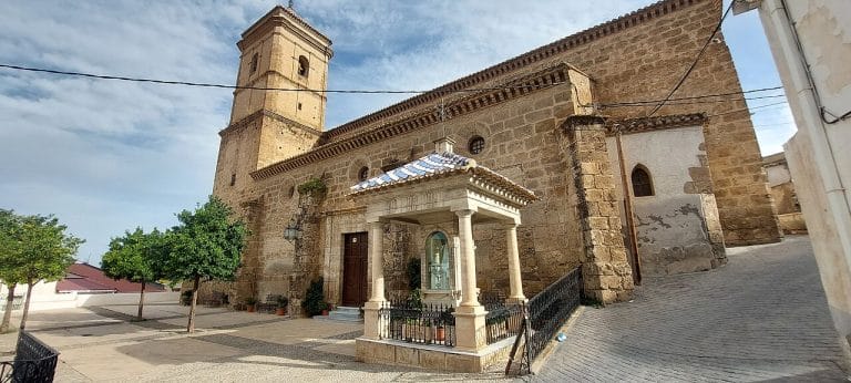 Vista frontal de la iglesia de San Ginés en Purchena, con muros de sillería envejecida y una torre de campanario a la izquierda; en primer plano, un templete con tejado de tejas azules que cobija una imagen religiosa, rodeado de una barandilla metálica, y a su alrededor, árboles y pavimento empedrado bajo un cielo parcialmente nublado.