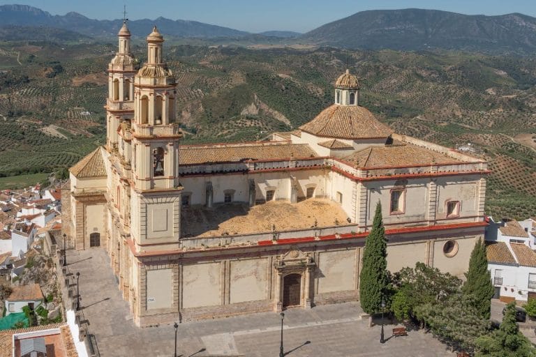 Fachada de la iglesia de Nuestra Señora de la Encarnación en Olvera (Cádiz) con sus dos torres barrocas y paisaje de sierras andaluzas al fondo.