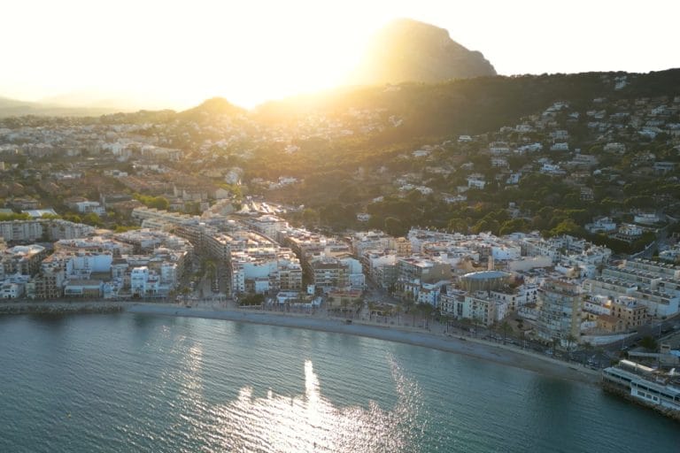 Vista aérea de una cala en Jávea, con formaciones rocosas, arena clara y el mar turquesa rompiendo en la orilla