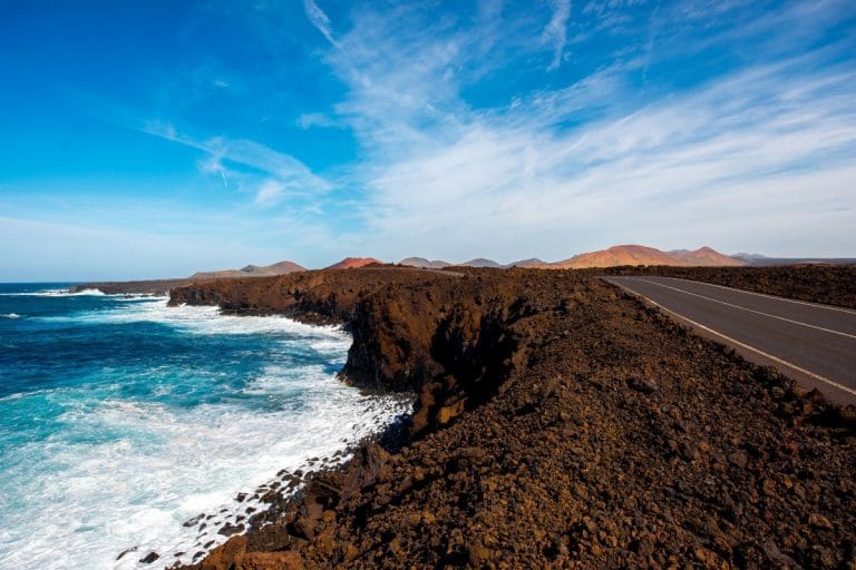 Acantilados volcánicos de Los Hervideros en Lanzarote con carretera junto al mar y montañas al fondo