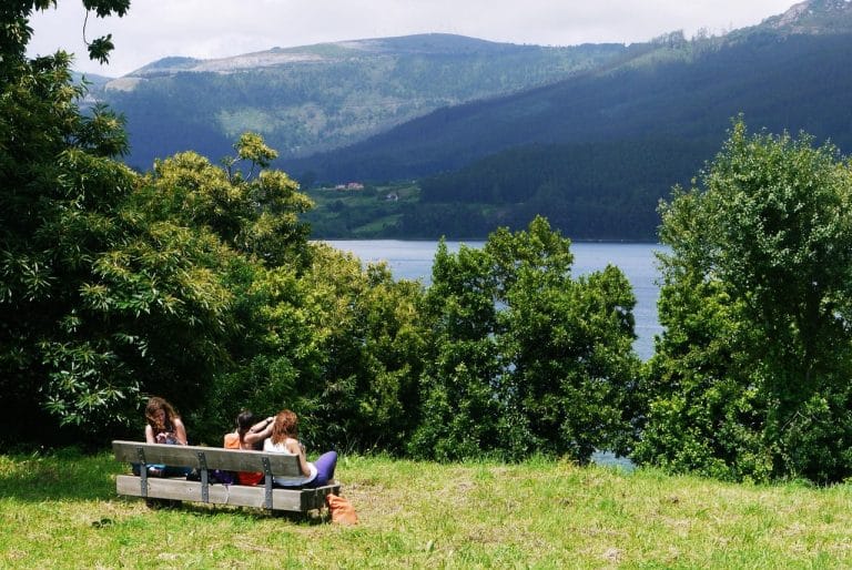 Tres personas sentadas en un banco de madera contemplan el embalse del río Navia, rodeado de bosques y montañas en un entorno natural de gran belleza.