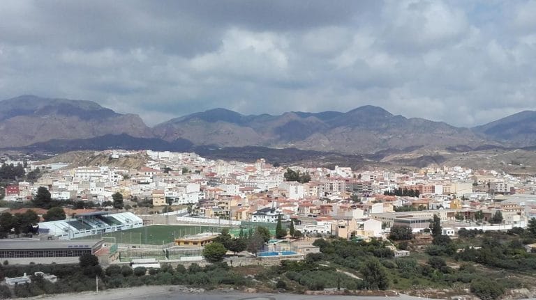 Vista panorámica de la localidad de Olula del Río, con edificios de fachada clara y tejados rojos, campos y vegetación en primer plano, y montañas en el horizonte bajo un cielo parcialmente nublado.