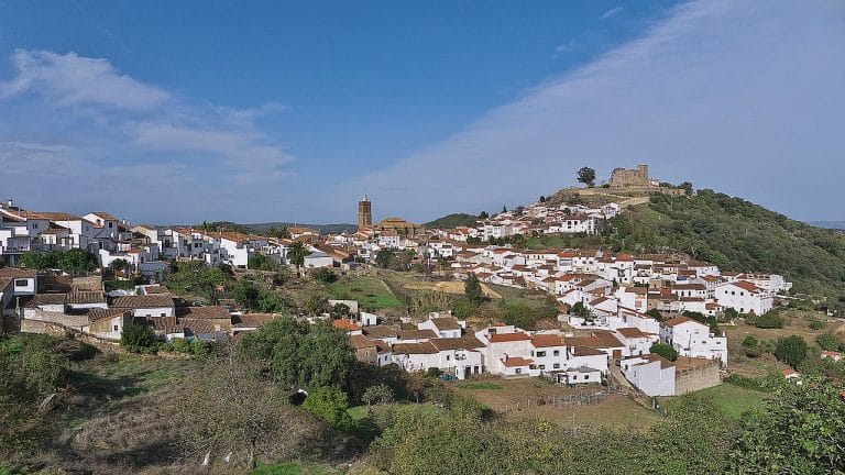 Vista panorámica del pueblo de Cortegana en Huelva, con sus casas blancas tradicionales y el castillo en lo alto de la colina rodeado de vegetación.