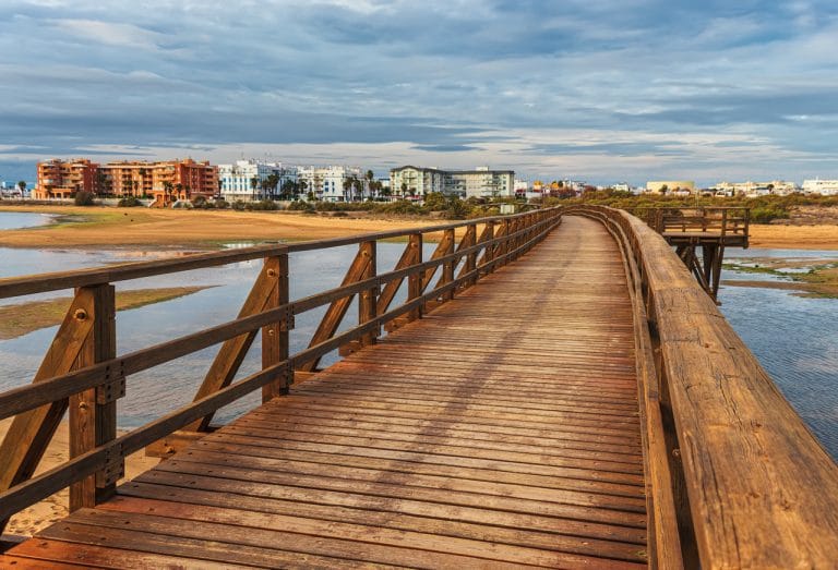 Pasarela de madera sobre la marisma al atardecer en Isla Cristina, con edificios al fondo y cielo parcialmente nublado.