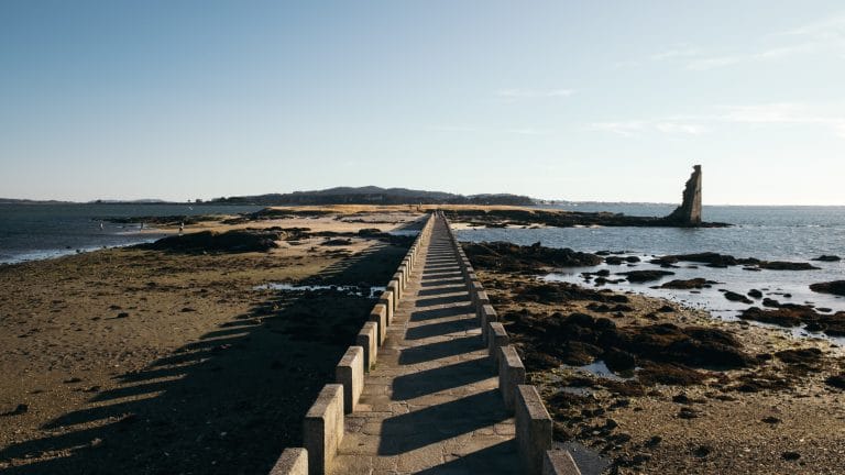 Larga pasarela peatonal de piedra sobre el mar, con la Torre de San Saturnino y la costa al fondo en la Isla de Arousa