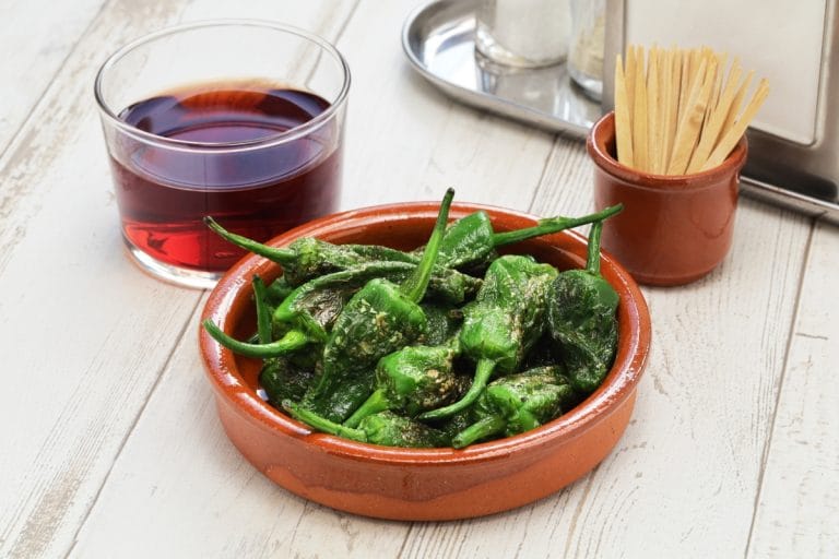 Plato de pimientos de Padrón asados con sal gruesa, acompañado de una copa de vino tinto, sobre mesa de madera clara.