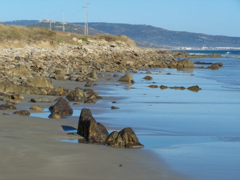 Vista de una playa con arena húmeda, rocas dispersas y suaves olas lamiendo la orilla, con colinas y líneas eléctricas en el horizonte