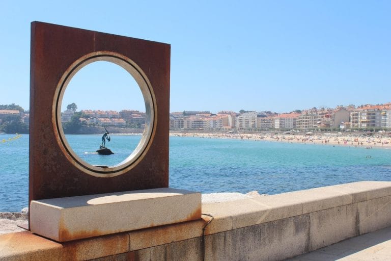 La icónica playa de Silgar en Sanxenxo, Pontevedra, con la escultura del Sireno en primer plano y la arena llena de bañistas bajo un cielo azul.