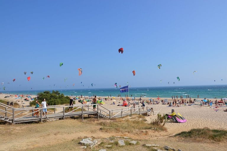 Playa de Valdevaqueros en Tarifa con numerosos kitesurfistas y bañistas disfrutando del sol, y pasarelas de madera en la arena.