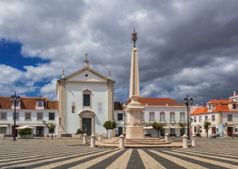 Vista de la Praça do Marquês de Pombal en Vila Real de Santo António, Portugal, con su iglesia, obelisco central y suelo empedrado en blanco y negro.