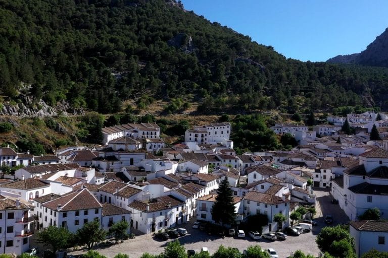 Panorámica de Grazalema (Cádiz) con casas blancas apiñadas en la base de una ladera arbolada y montañas al fondo.