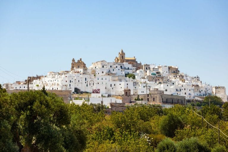Vista panorámica de un pueblo blanco con casas encaladas dispuestas en una colina, rodeado de vegetación y con dos torres de iglesia en la parte más alta.