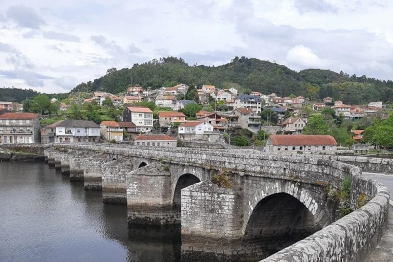 Vista del puente medieval de Arcade sobre el río Verdugo, con el pueblo al fondo y colinas boscosas en el horizonte.