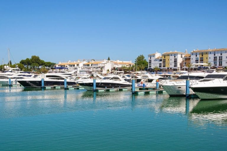 Yates de lujo atracados en el puerto deportivo de Vilamoura con edificios coloridos al fondo y cielo despejado.