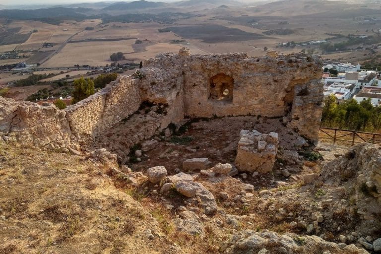 Ruinas de un antiguo castillo en lo alto de una colina con muros de piedra erosionados, rodeadas de terreno árido; al fondo se observan campos agrícolas y un pequeño pueblo de casas blancas bajo un cielo despejado.