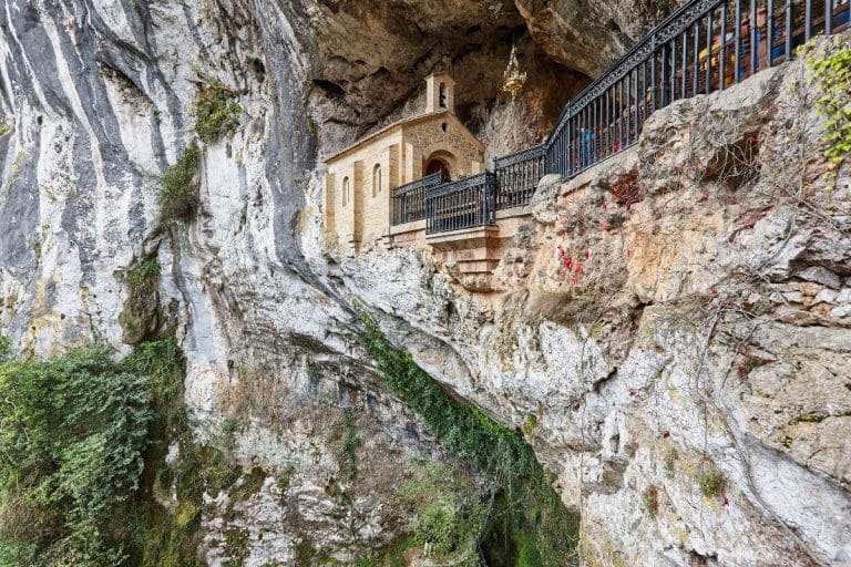Capilla de la Santa Cueva de Covadonga encajada en un acantilado de roca caliza, con barandilla de hierro forjado y vegetación colgante