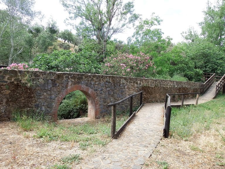 Sendero empedrado con pasamanos de madera en Puerto Moral, atravesando un entorno verde con flores, árboles y un antiguo puente de piedra y ladrillo