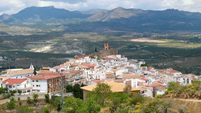 Vista panorámica del pueblo de Serón en Almería, con sus casas de tejados rojos y muros blancos asentadas en la ladera; al fondo se aprecian campos de cultivo y montañas bajo un cielo parcialmente nublado.