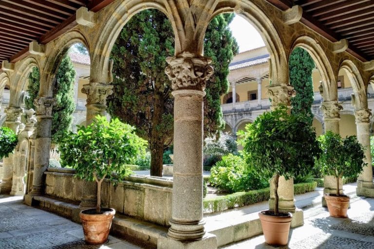 Claustro del Monasterio de Yuste en Cáceres, con columnas de piedra, arcos góticos y jardín central con cipreses y arbustos.