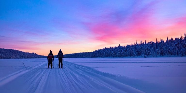 Dos personas esquiando sobre un lago congelado en Torassieppi, Finlandia, al atardecer con cielo rosado