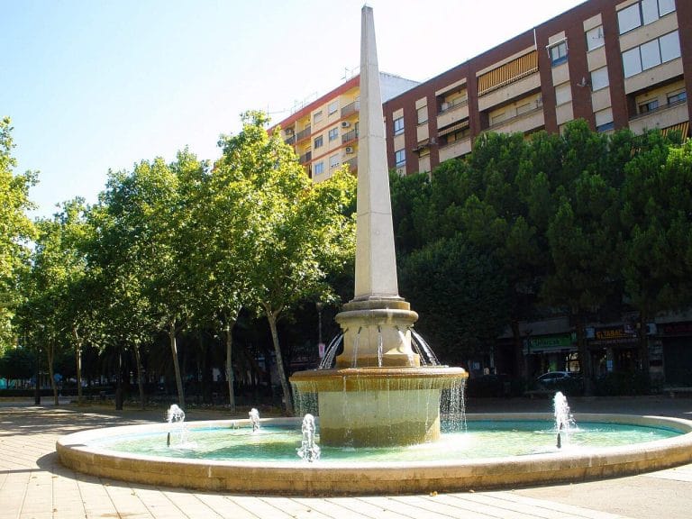 Fuente con obelisco en el Paseo de San Gregorio de Puertollano, rodeada de árboles y edificios