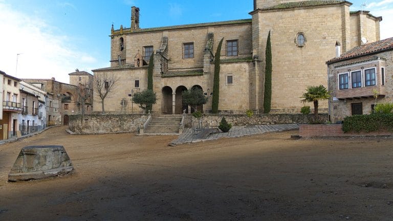 Fachada principal de la iglesia de Nuestra Señora de la Asunción en Oropesa, Toledo, con árboles sin hojas y plaza adoquinada en primer plano