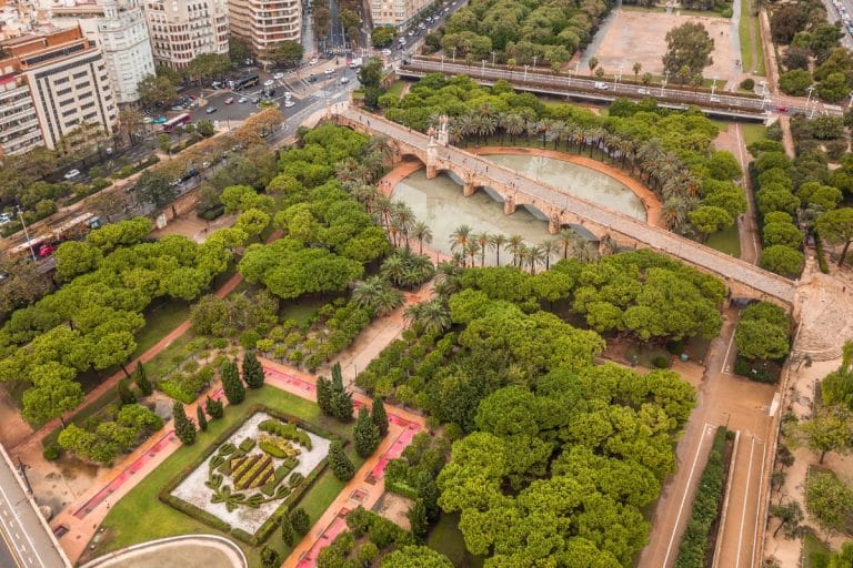 Vista aérea del Jardín del Turia en Valencia, mostrando sus extensas zonas verdes, un puente arqueado, palmeras y edificios urbanos en el horizonte.
