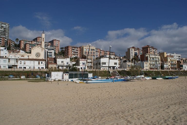 Playa de Montgat con embarcaciones varadas en la arena y casas frente al mar bajo un cielo azul con nubes