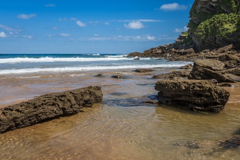 Vista de la playa de Zarautz con formaciones rocosas y olas del mar Cantábrico en un día soleado.